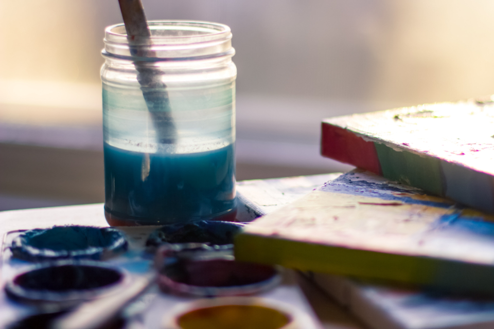 A jar of water with a paintbrush beside a well-used watercolour palette